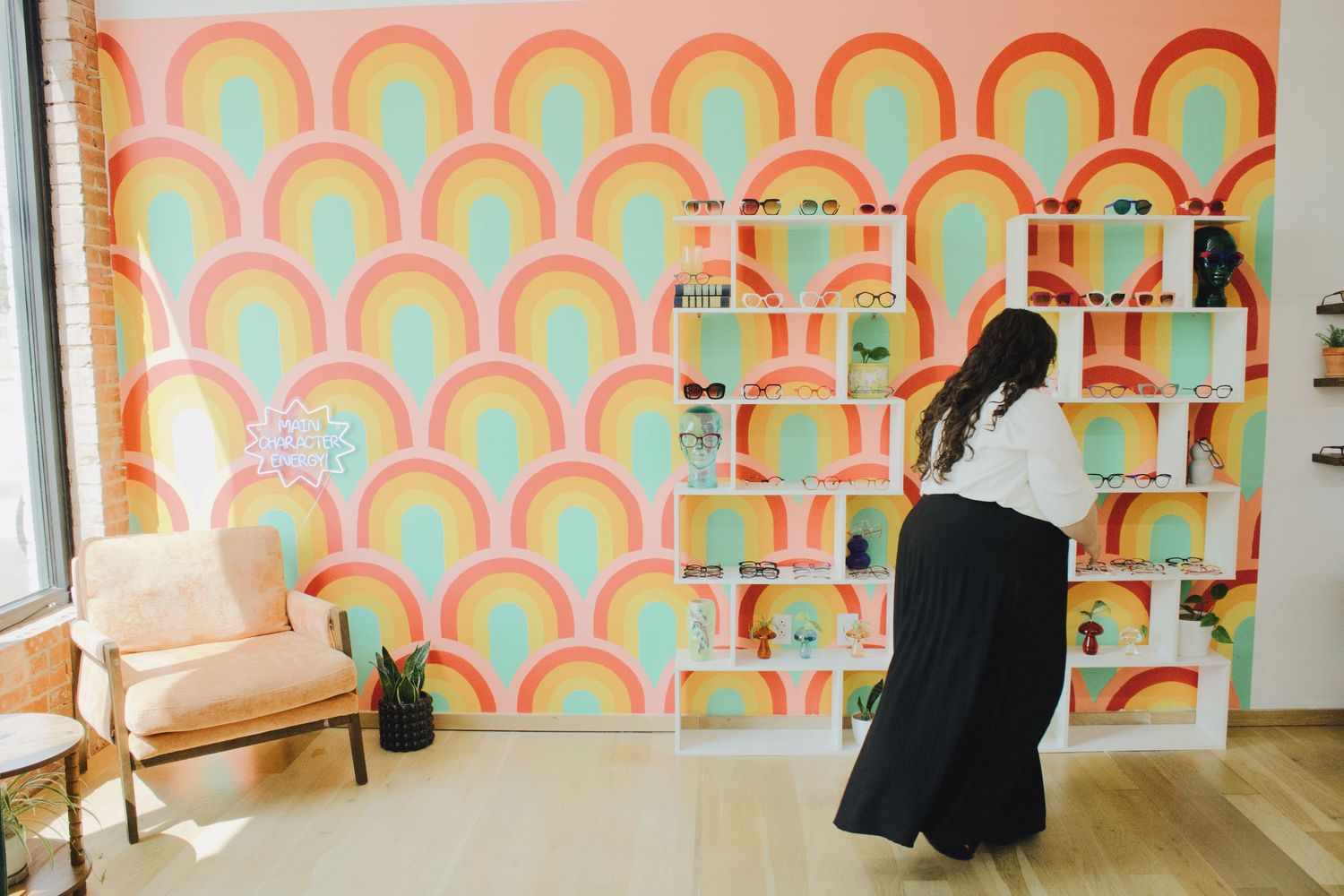 Woman browsing white shelving unit displaying eyeglasses against a colorful rainbow mural wall.