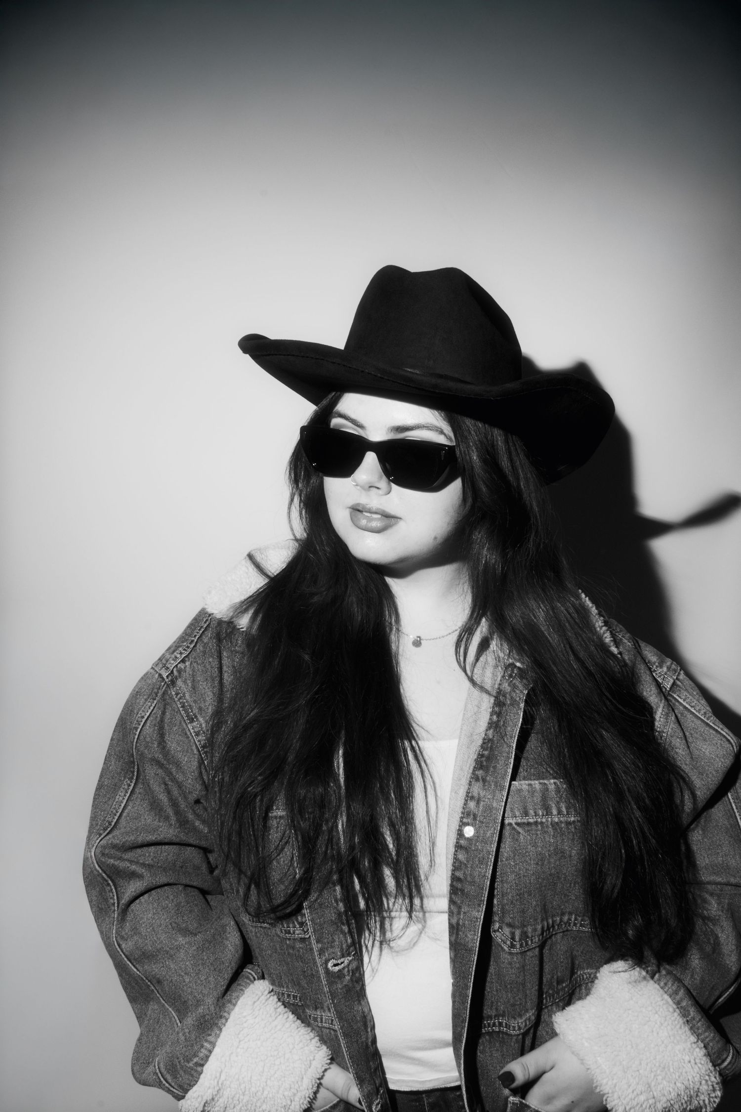 Black and white portrait of woman in cowboy hat, sunglasses, and denim jacket posing against wall.