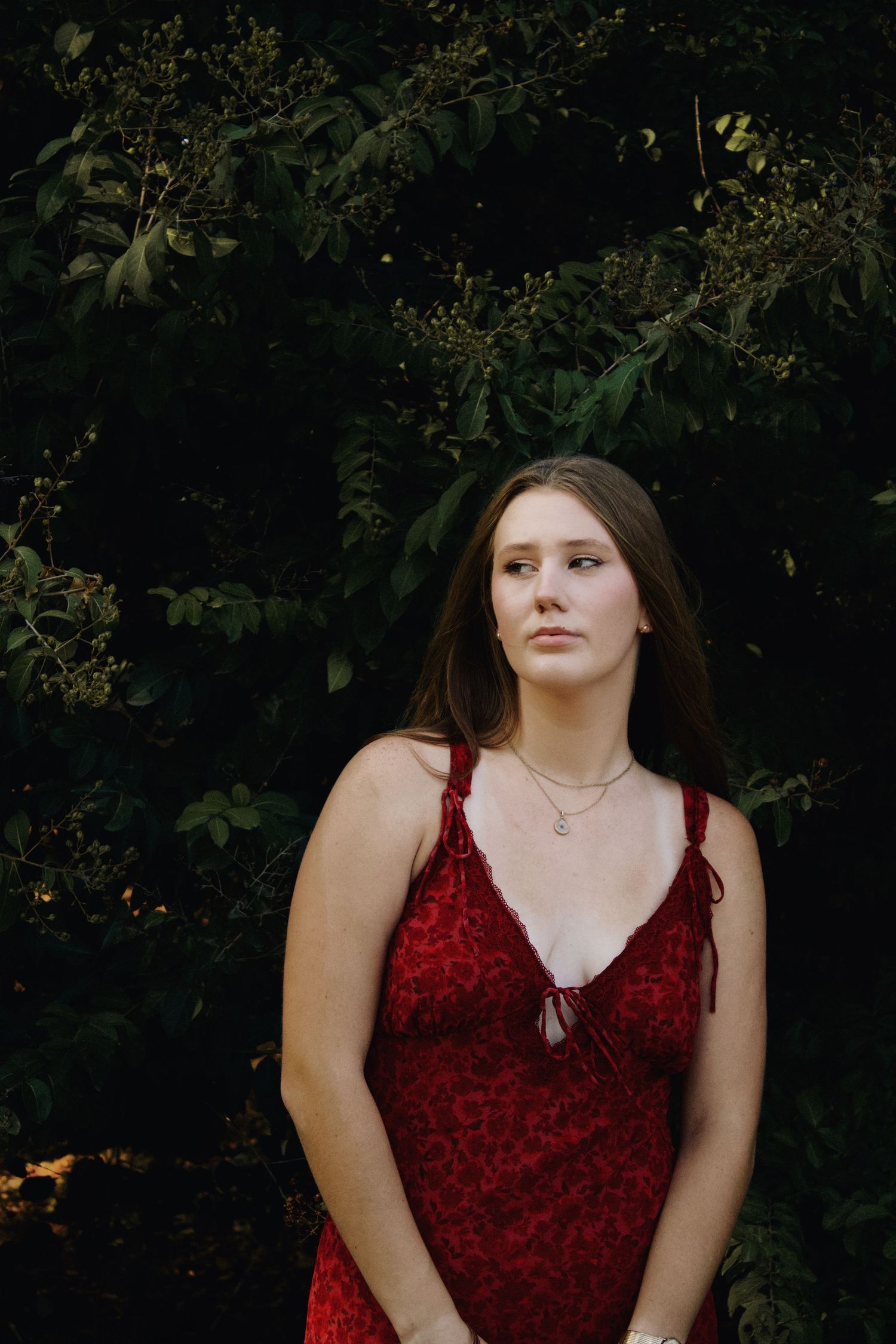 Young woman in red floral dress standing before lush dark green foliage outdoors.