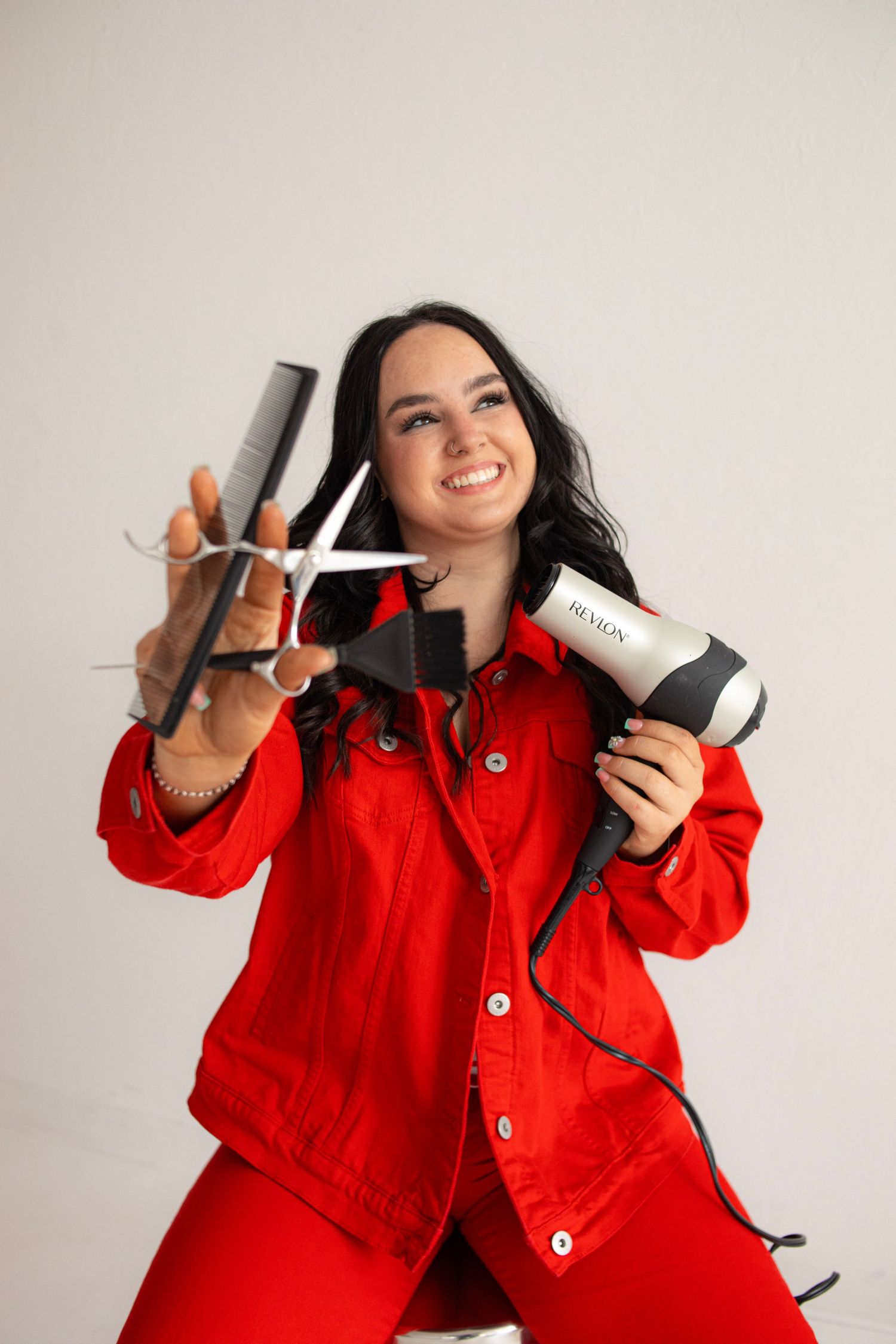 Smiling female hairstylist in red outfit holding Revlon hair dryer and scissors with comb against white background.