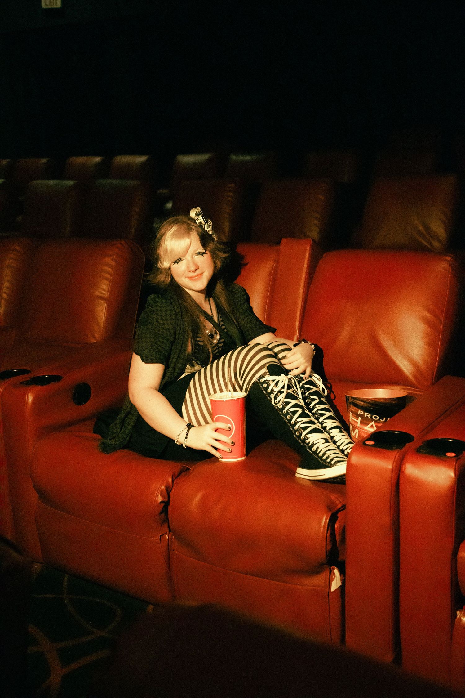 A young woman in striped tights and platform sneakers relaxes in red leather cinema seats holding a drink.