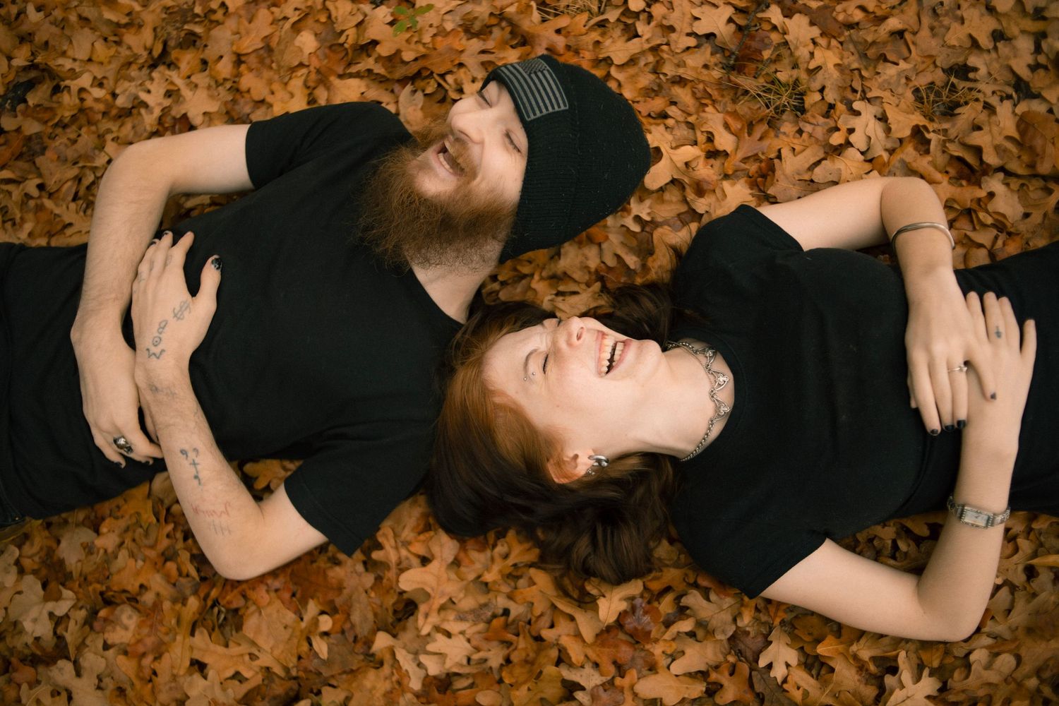 Couple in black shirts laughing while lying on autumn leaves outdoors.