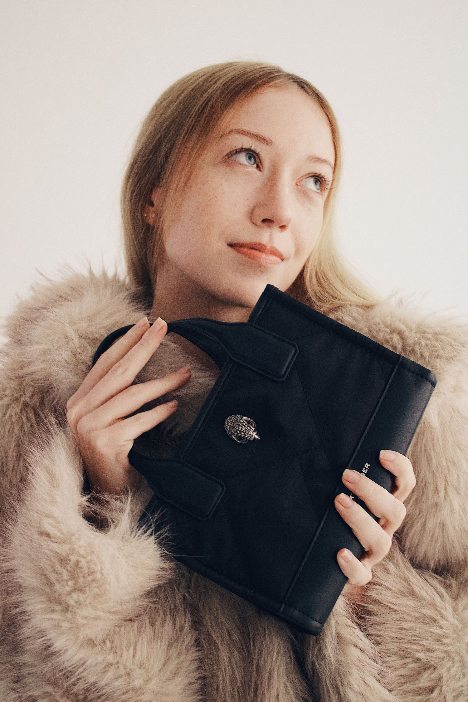 Blonde woman in faux fur coat holding black quilted clutch bag, gazing upward thoughtfully.