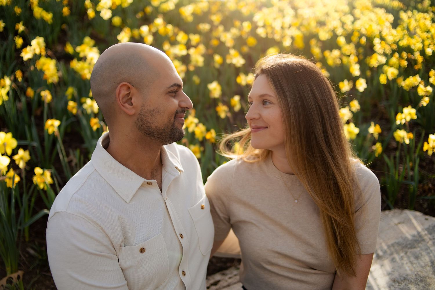 Couple smiling at each other in front of blooming yellow daffodils in golden sunlight.