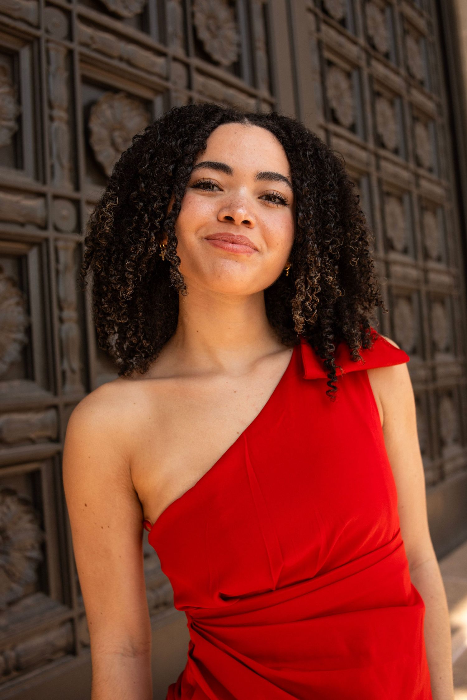 Young woman with curly hair smiling in red one-shoulder dress against ornate door.