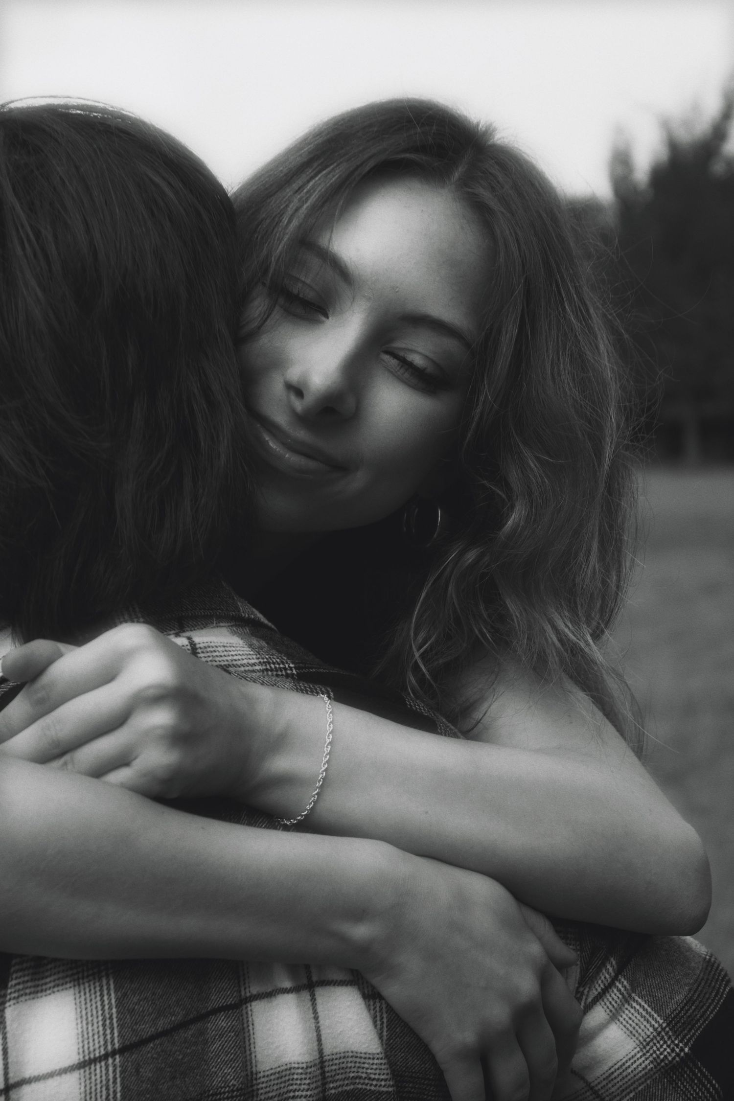 Black and white photo of a young woman smiling softly while hugging someone close outdoors.