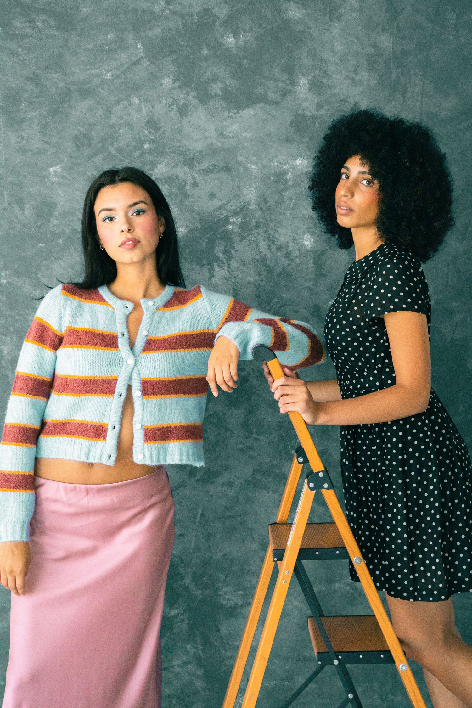 Two women pose against gray wall, one in striped cardigan and pink skirt, other in polka dot dress.