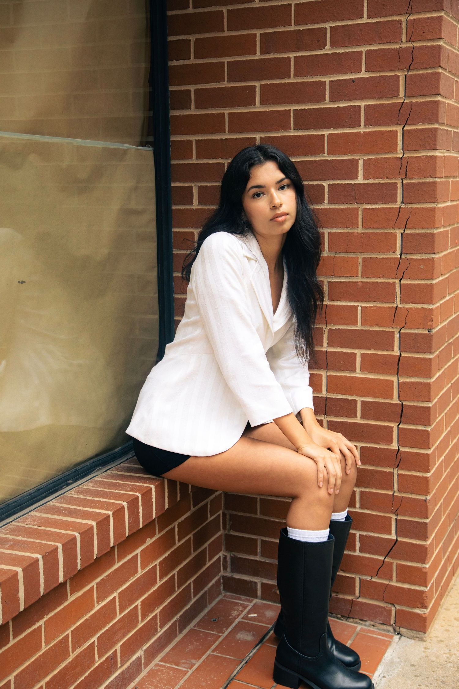 Young woman in oversized white shirt and black boots sitting on a brick window ledge.