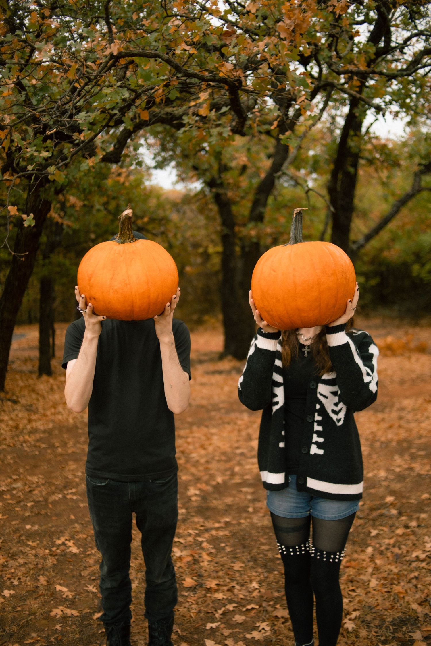 Couple holding large orange pumpkins in front of their faces in an autumn forest setting.