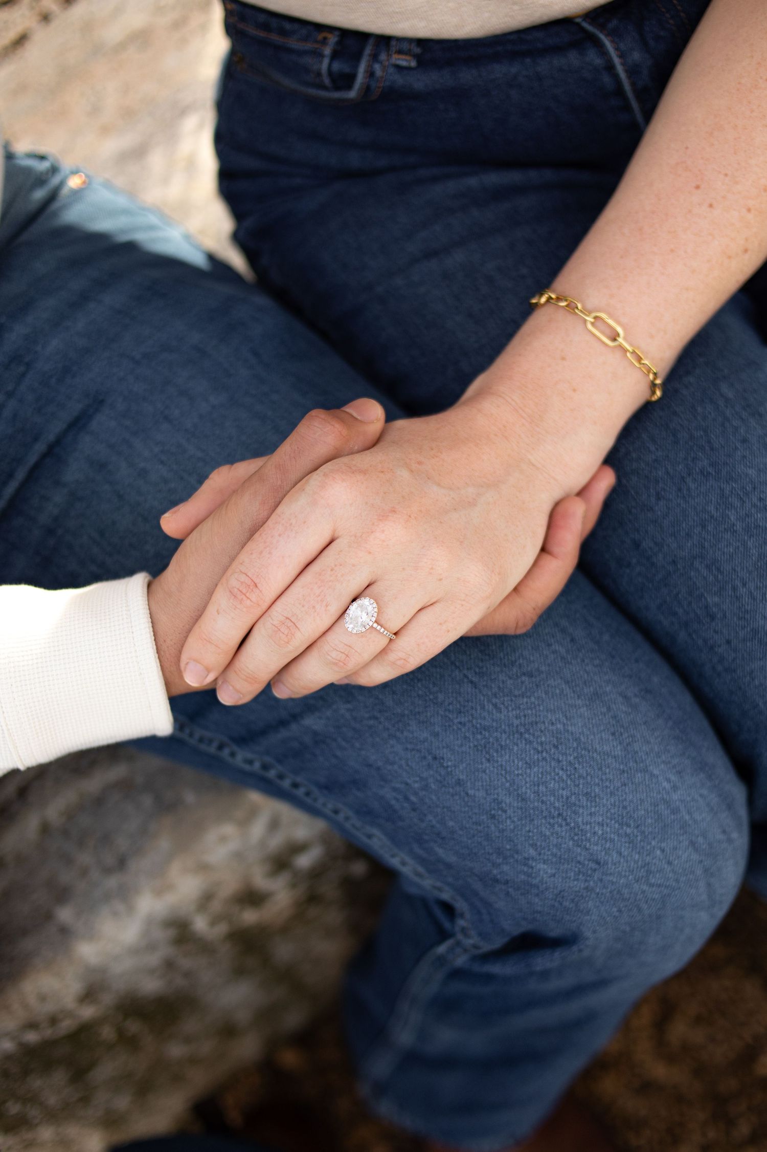 Close-up of couple holding hands showing oval diamond engagement ring and gold chain bracelet.