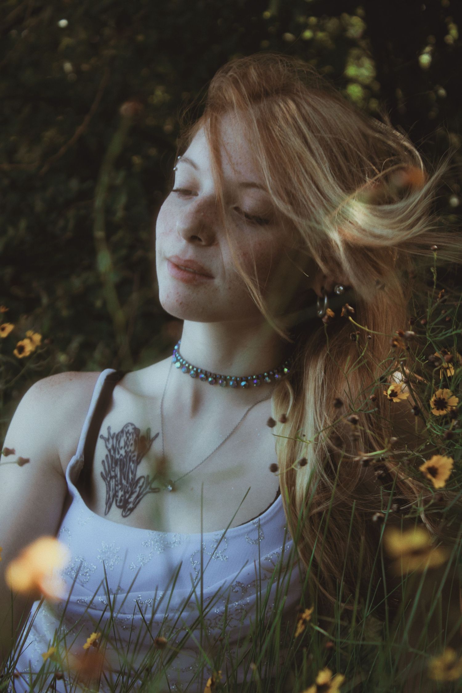 Young woman with blonde hair and chest tattoo sitting among yellow wildflowers with eyes closed.