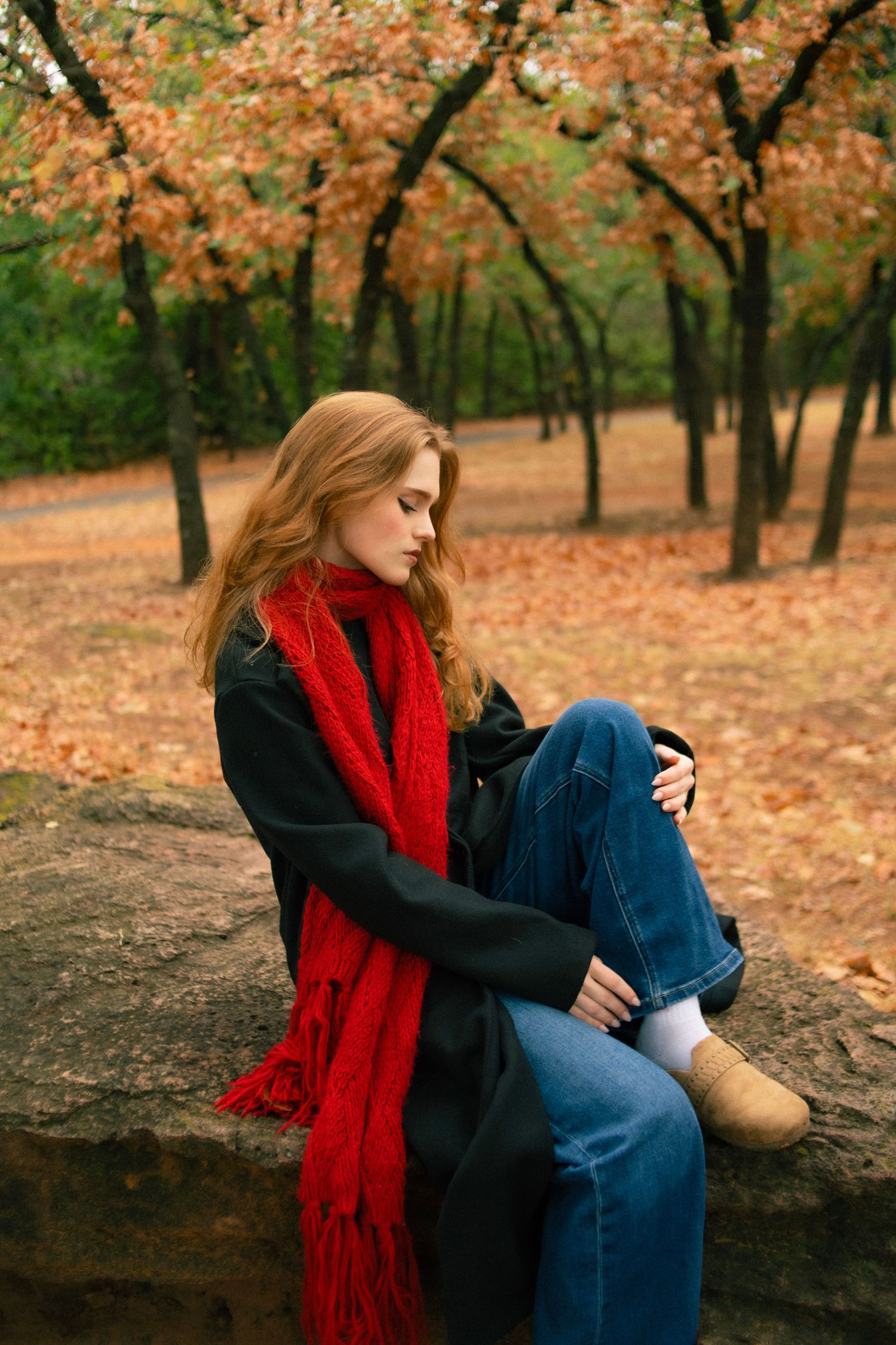 Young woman with red scarf and black coat sits on stone wall in autumn park with colorful foliage.