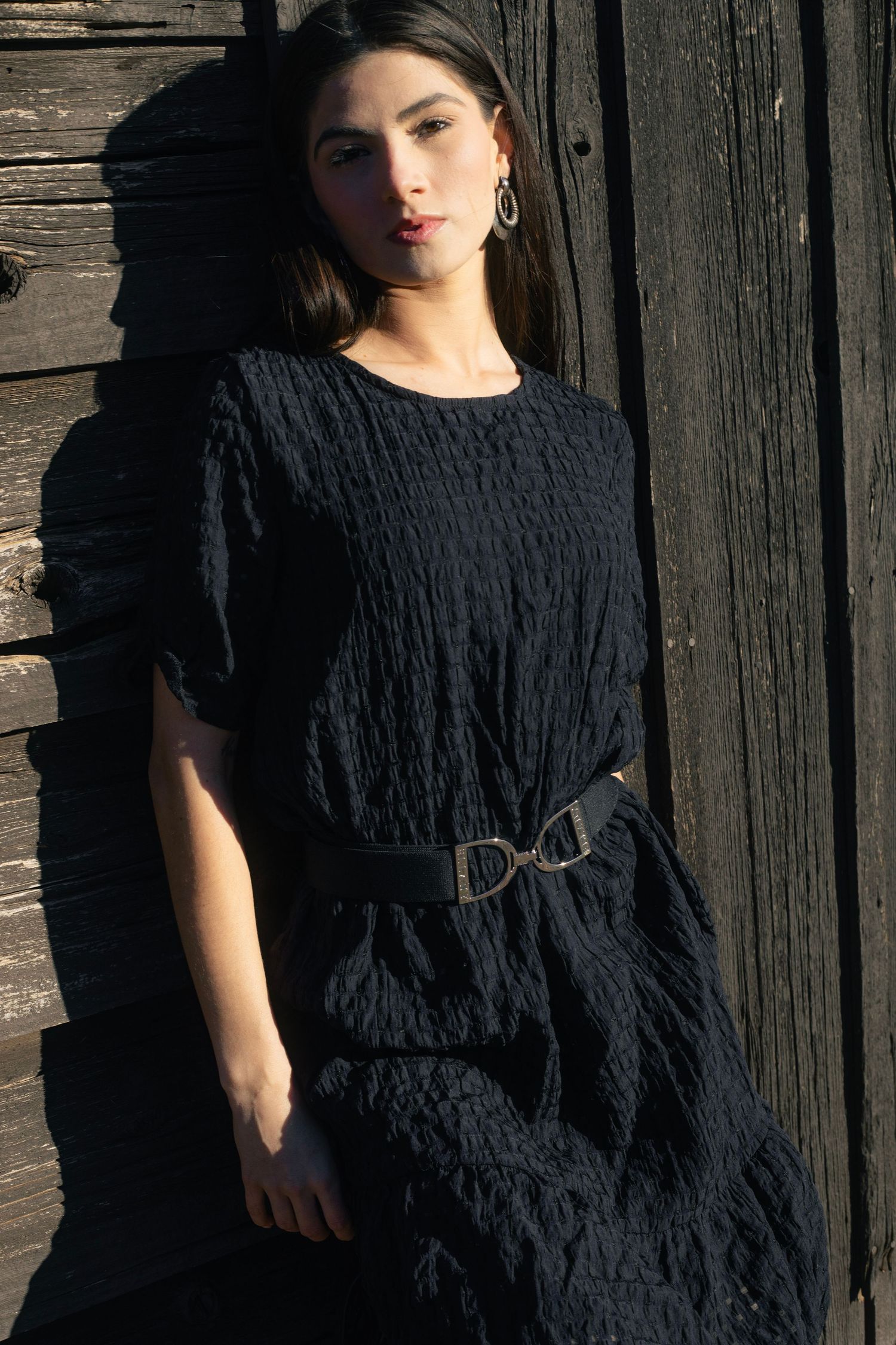 Woman in black textured dress with D-ring belt leaning against dark rustic wooden wall.