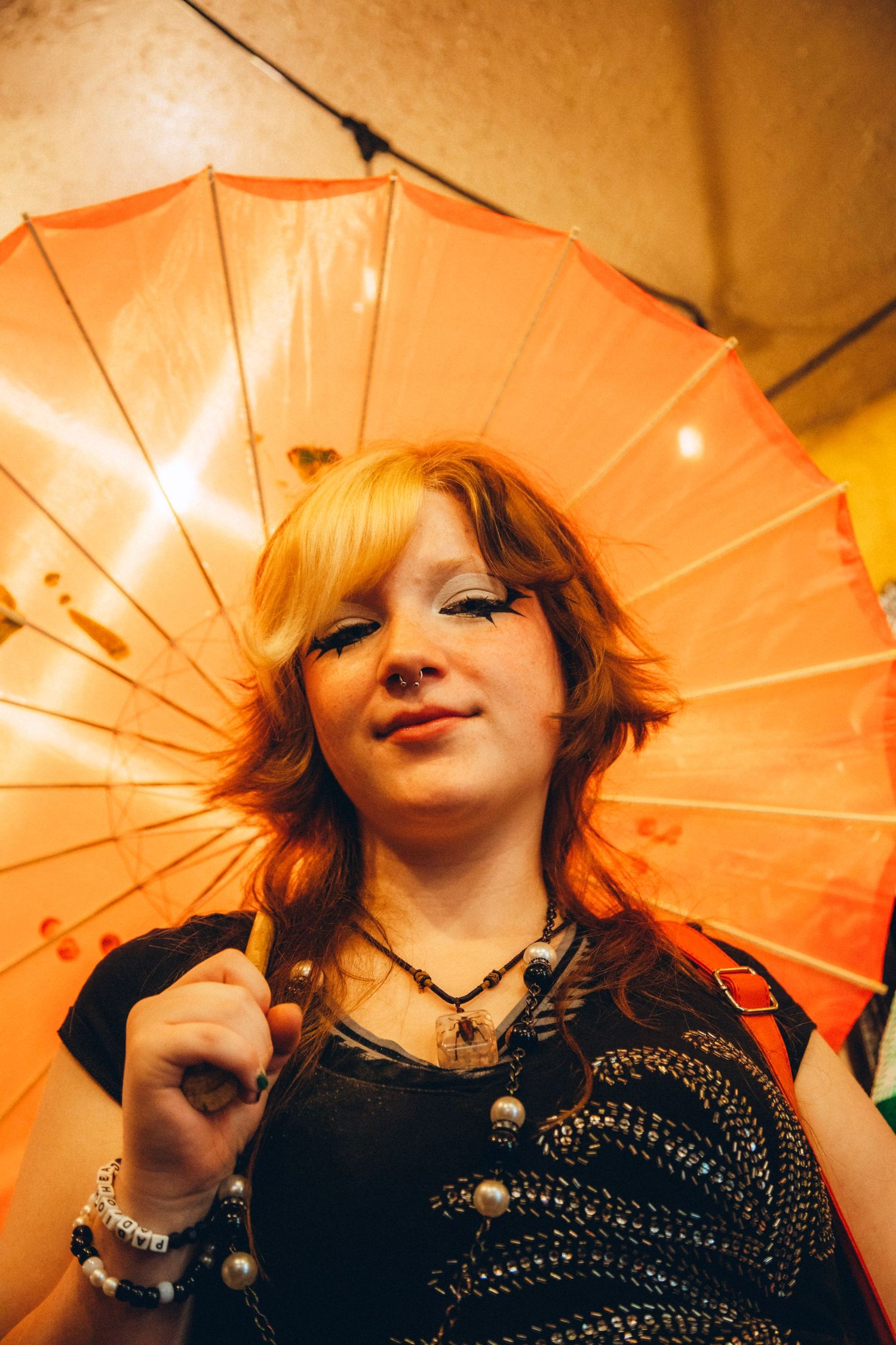 Young redhead woman holding an orange parasol umbrella with dramatic eye makeup indoors.