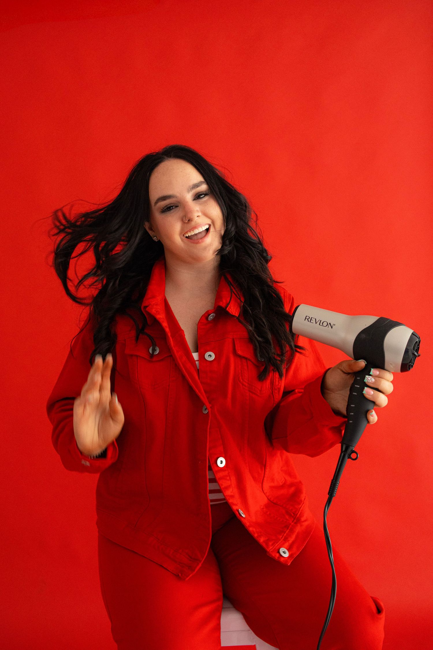 Woman in red outfit holding a Revlon hair dryer against a red background with flowing dark hair.