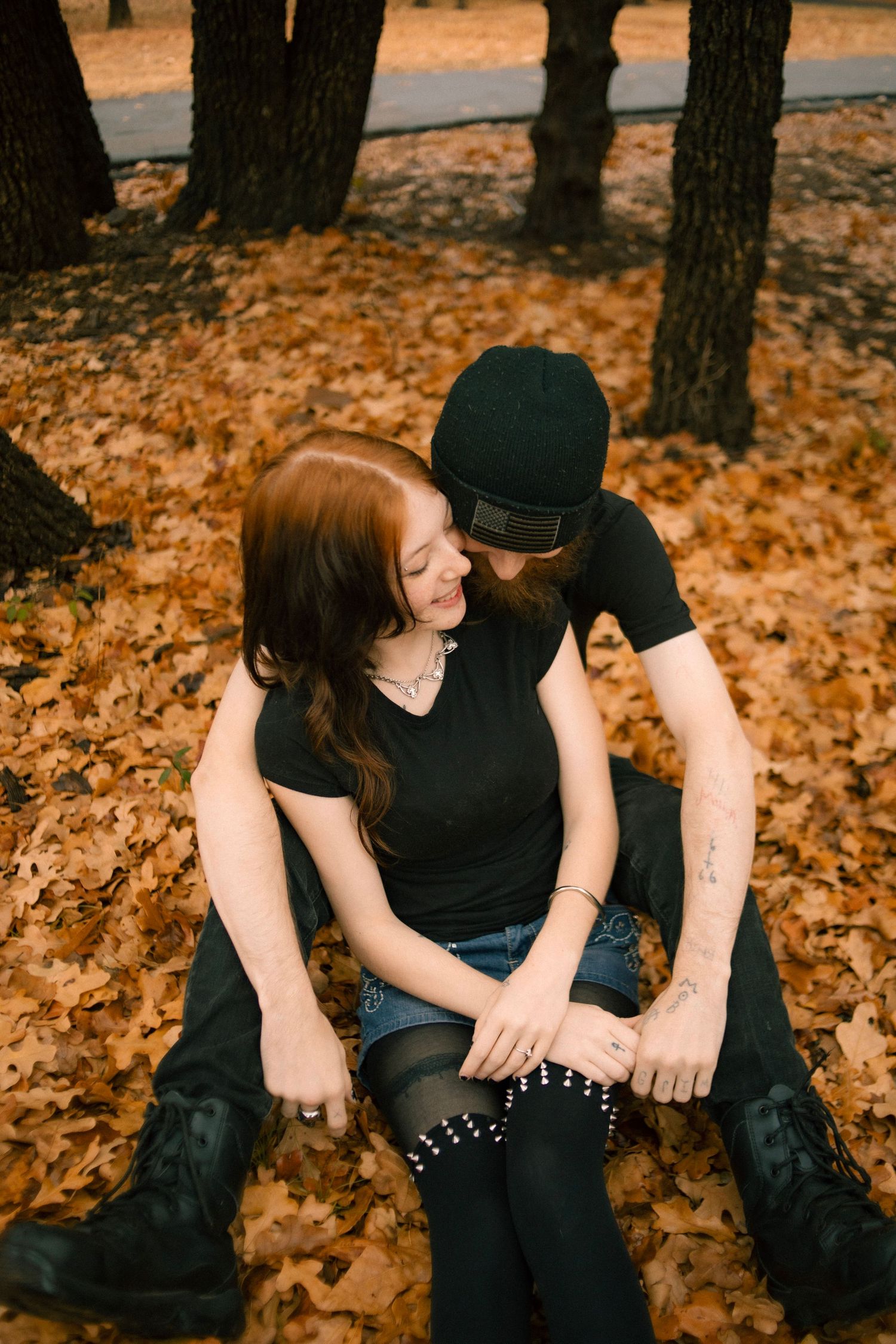 Couple sitting together on autumn leaves in a park, wearing black outfits, sharing a tender romantic moment.