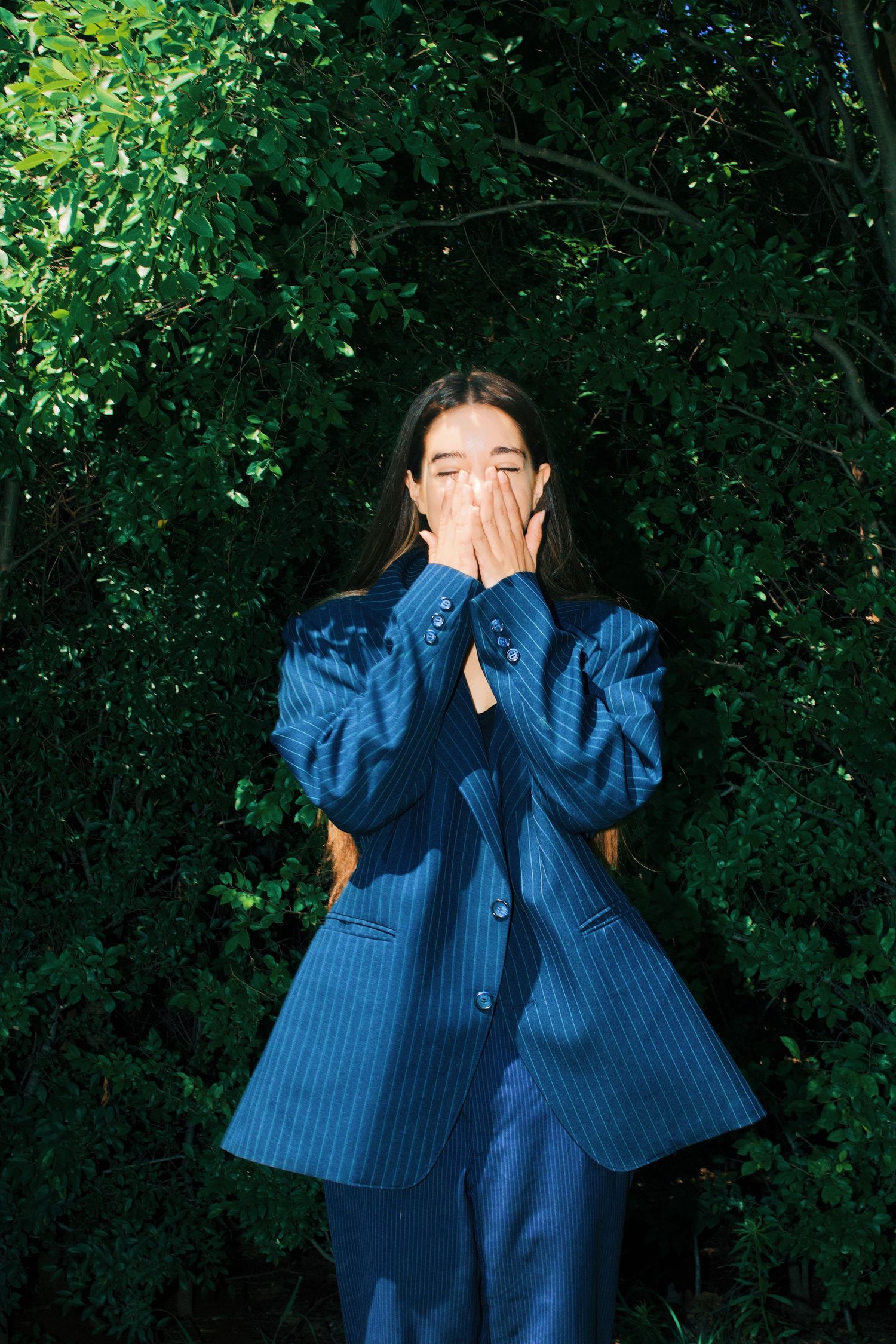Woman in blue pinstripe oversized suit covering her face with hands while standing in front of lush green bushes.