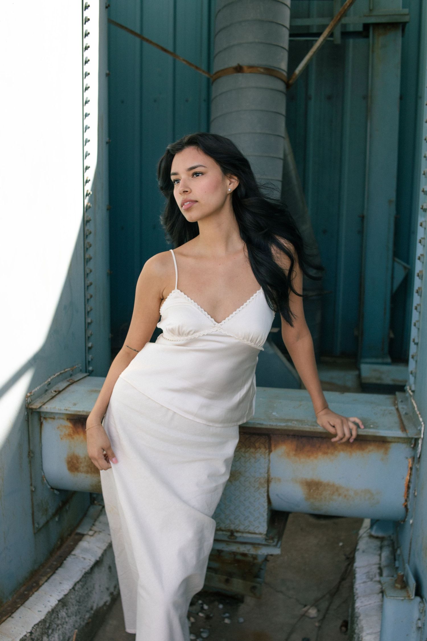 Young woman in white slip dress posing against rustic teal metal structure outdoors.