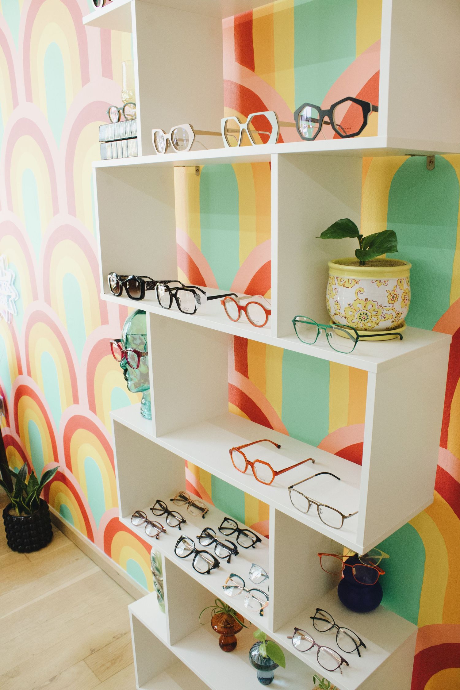White shelving unit displaying various eyeglass frames against a colorful rainbow patterned mural wall.