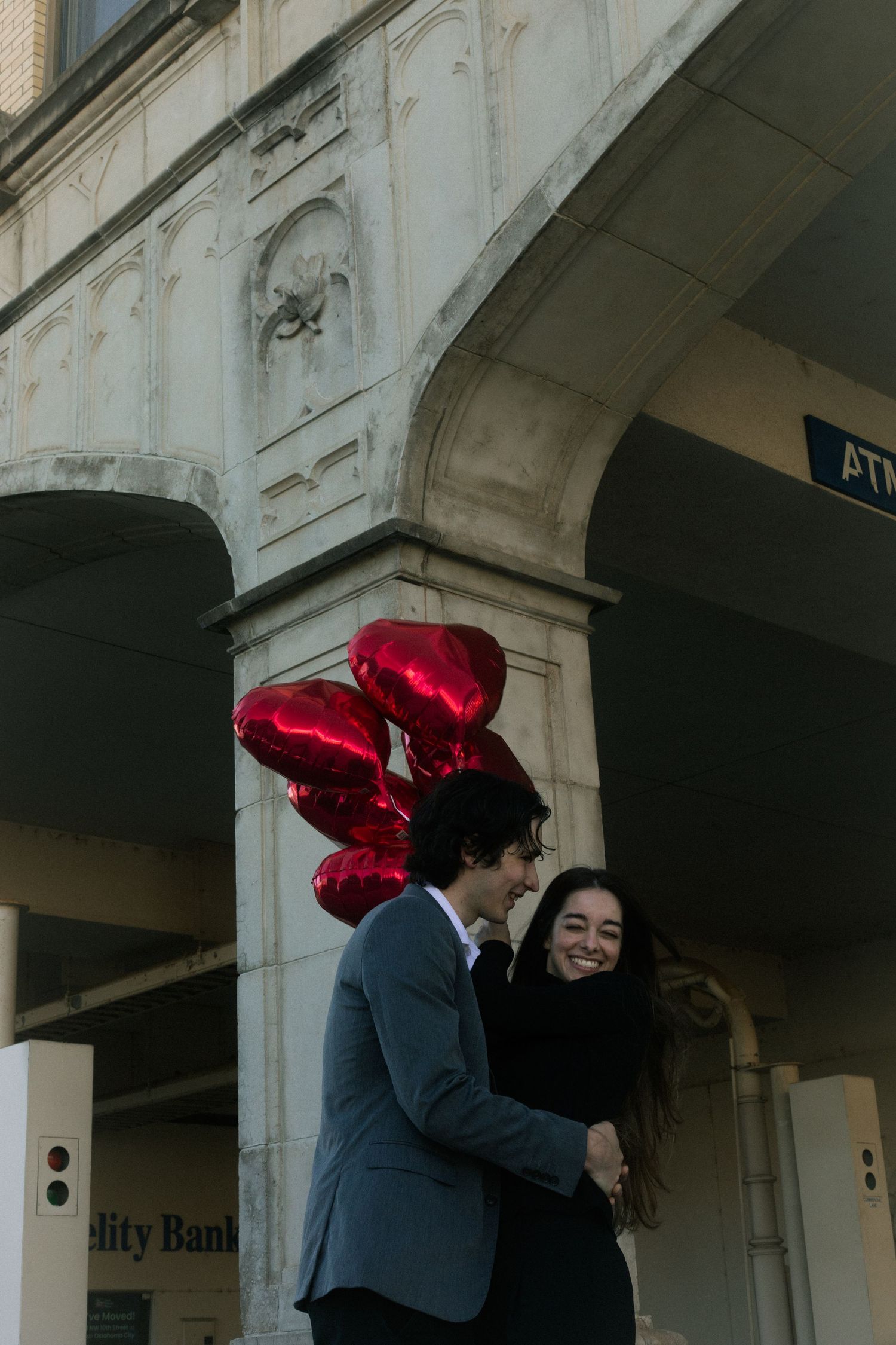 A joyful couple embraces under a stone archway, holding red heart-shaped balloons in an urban setting.