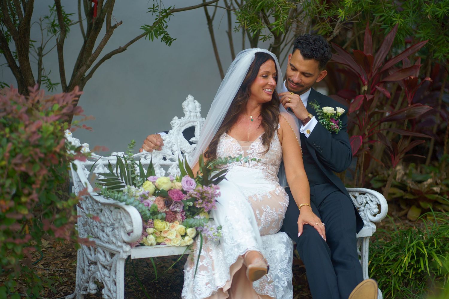 A romantic wedding couple sits together on an ornate white garden bench surrounded by lush greenery and flowers.