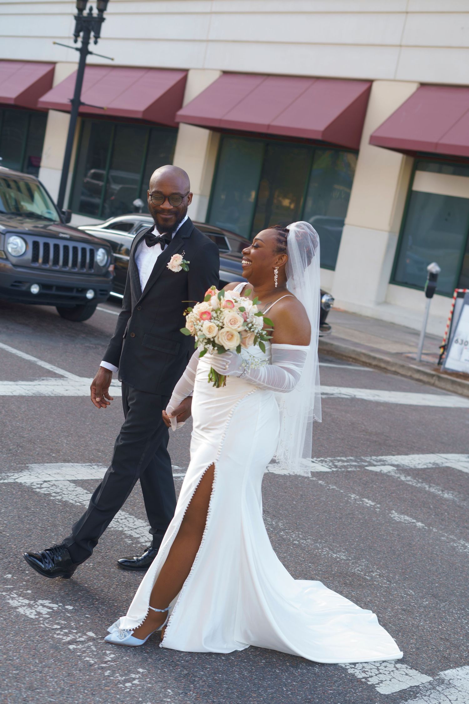Newlyweds walk across a street with bride in elegant white gown with slit holding peach bouquet and groom in black tuxedo.