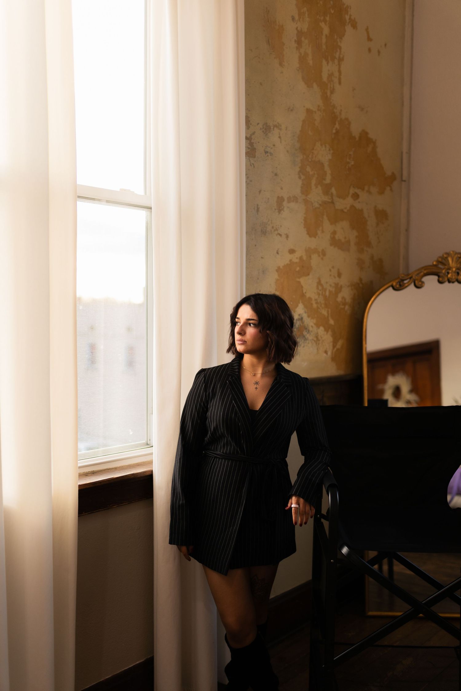 Person in black clothing leans against window frame in room with weathered walls and natural light streaming in.