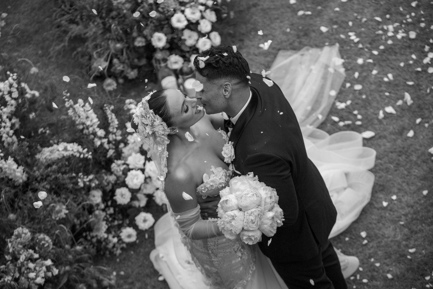 Black and white wedding photo of a romantic kiss surrounded by flowers and confetti.