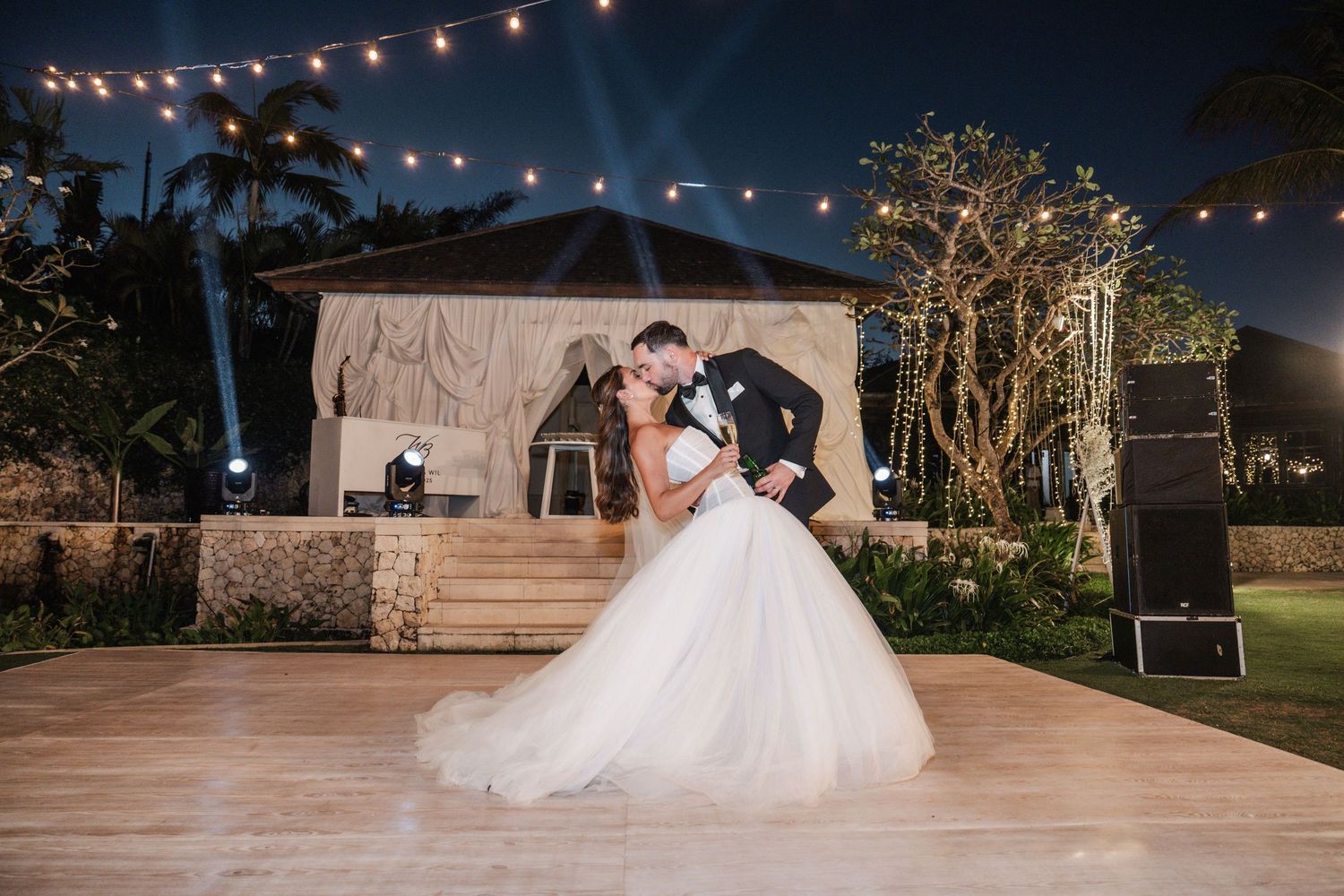 Newlywed couple shares romantic dance under string lights at outdoor evening wedding reception.