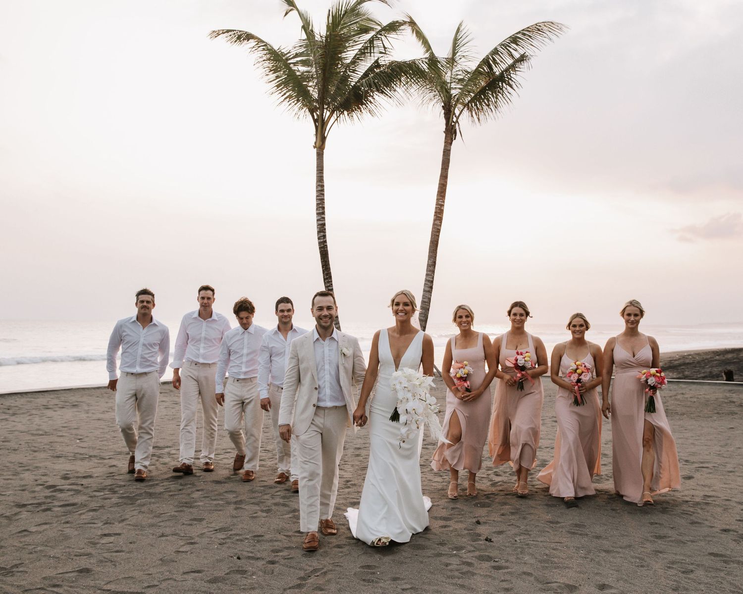 Tropical beach wedding party with bridesmaids in blush dresses and groomsmen in white suits by palm trees.