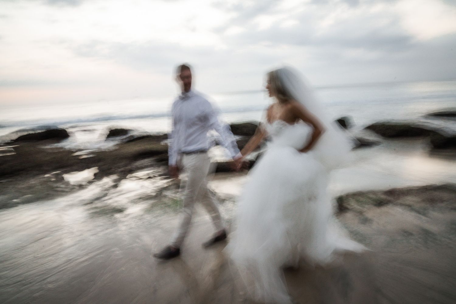 A couple in wedding attire runs along a beach at sunset creating a motion blur effect in the photograph.