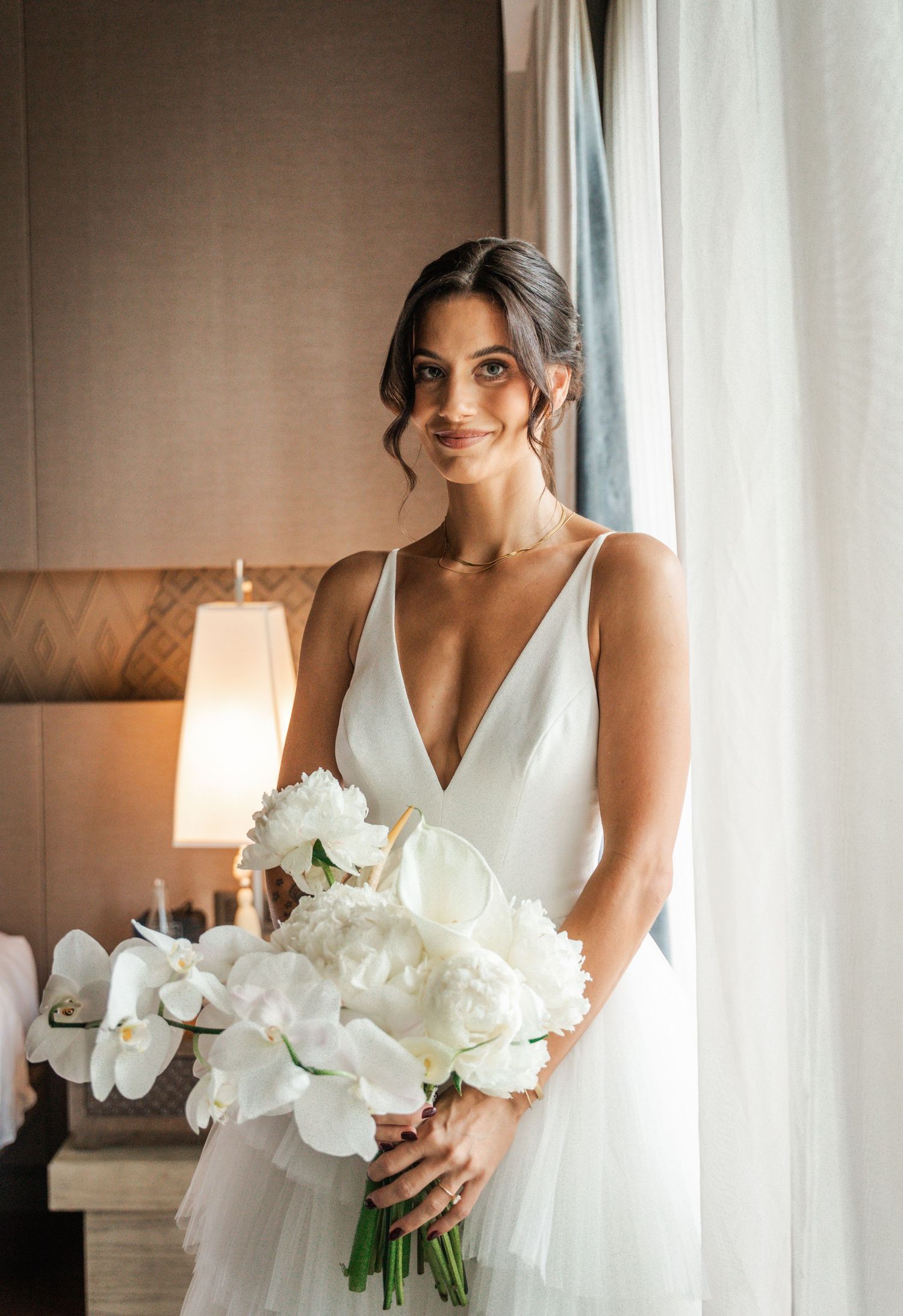 Bride in a deep V-neck white gown holds a white orchid and peony bouquet, standing by a window in soft natural light.