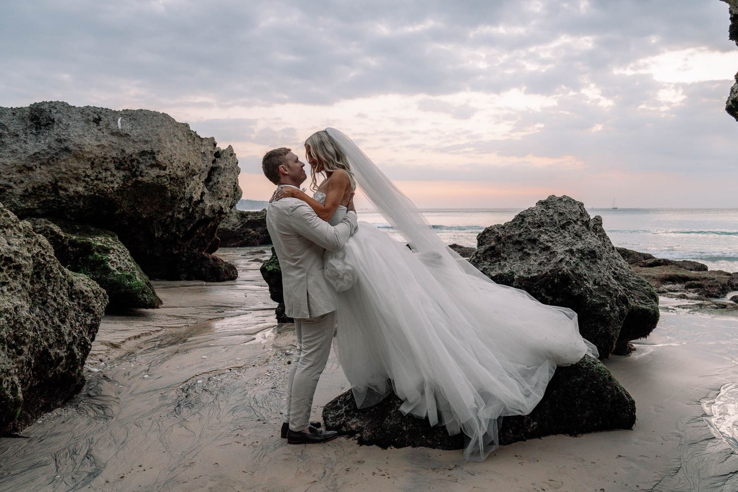 A couple in wedding attire poses romantically on a rocky beach at sunset with flowing white dress and dramatic sky.