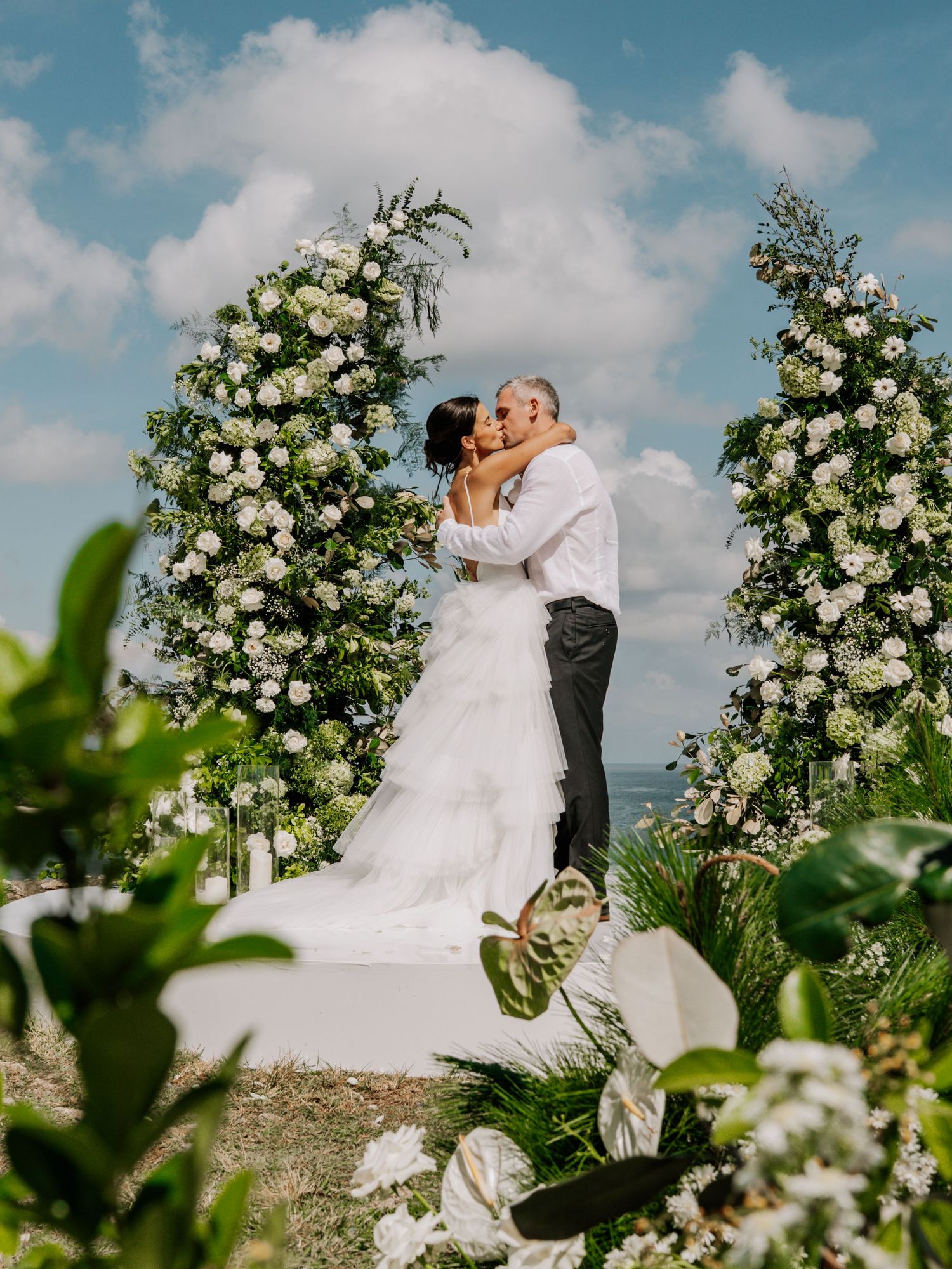 Bride and groom share first kiss at outdoor wedding altar adorned with white floral arrangements under a blue sky.