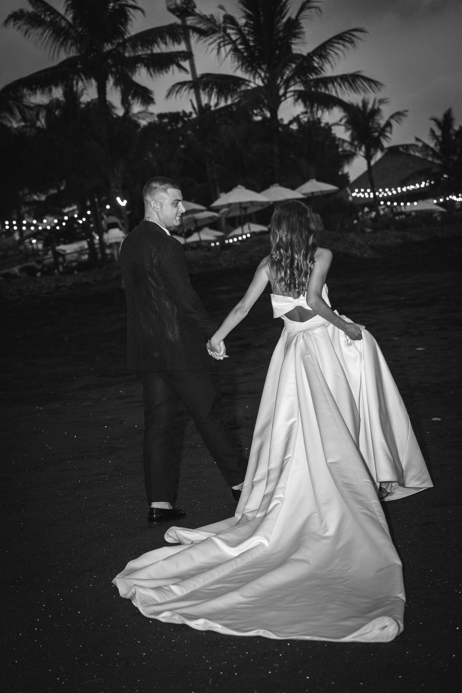 A couple dances at a romantic outdoor wedding reception with palm trees and string lights in black and white.