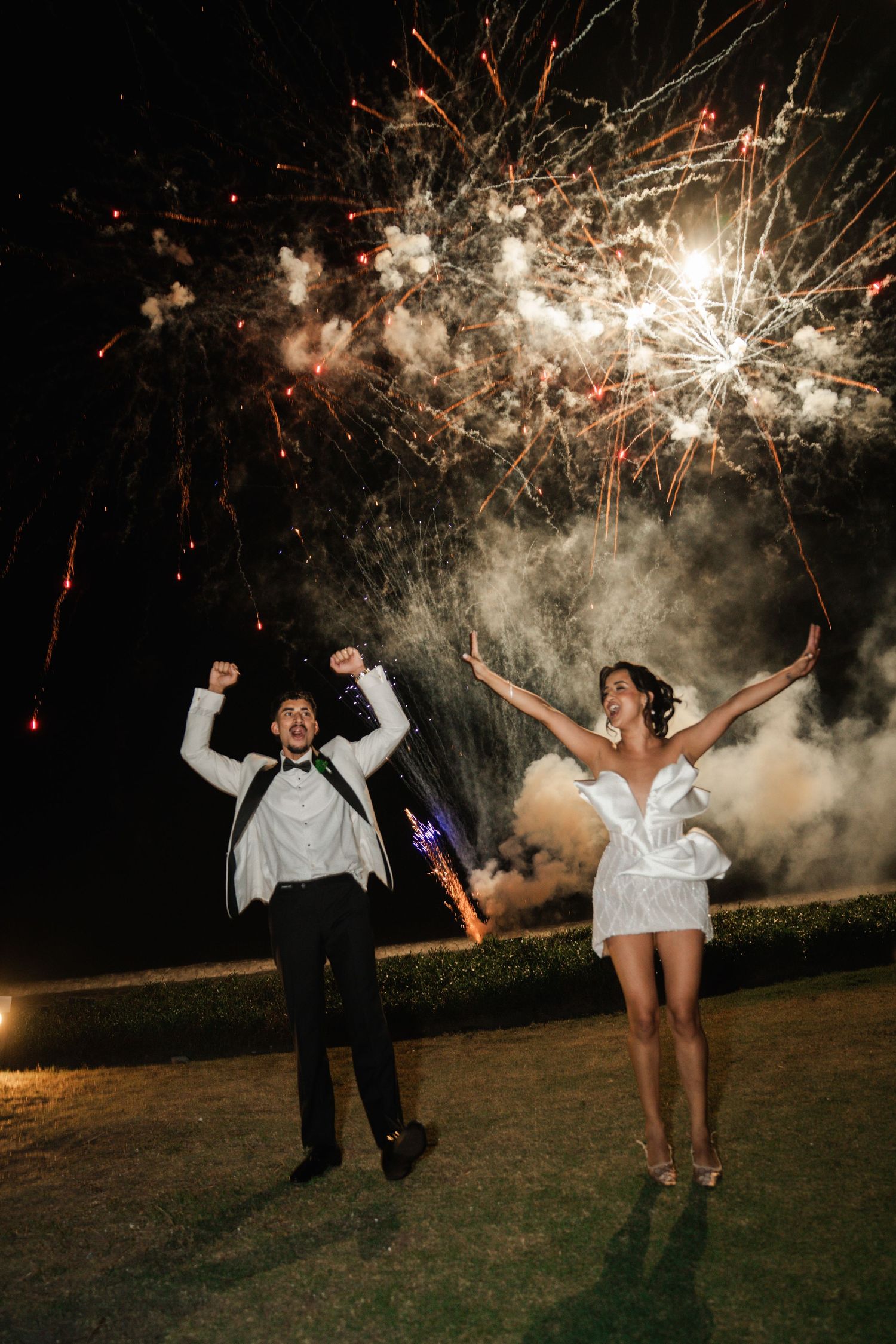 A couple celebrates with joyful jumping poses under spectacular fireworks display at an outdoor nighttime celebration.