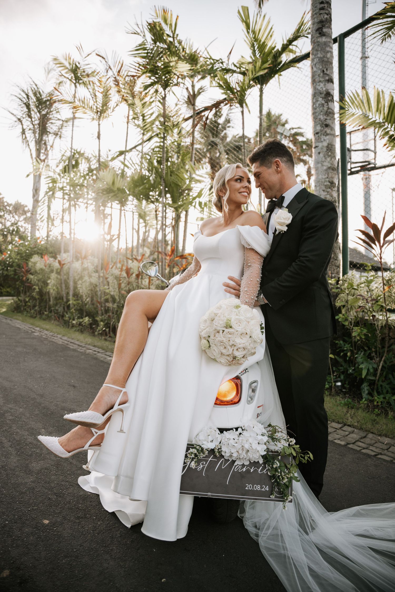 A couple in black suit and white wedding dress share intimate poses along a palm-lined road at sunset in Bali