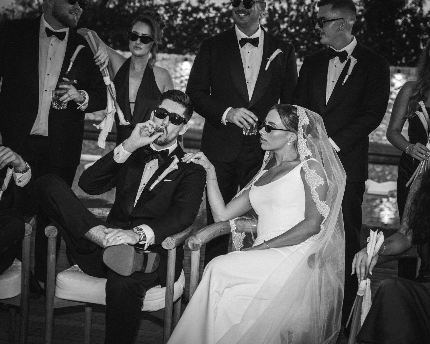 Black and white wedding photo of bride and groom in sunglasses sharing a drink with wedding party outdoors.
