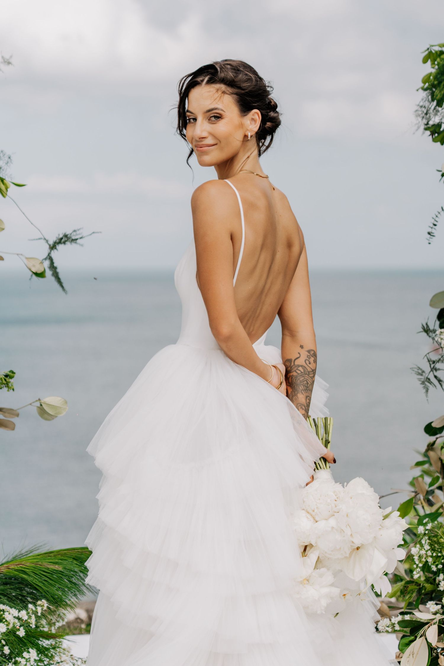 Smiling bride in backless white tulle gown holds peony bouquet at oceanside wedding ceremony.
