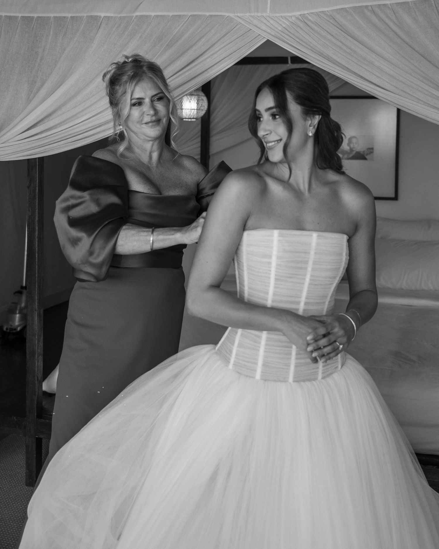 Black and white photo of two women getting ready in a bedroom, one wearing a wedding gown and one in formal dress.