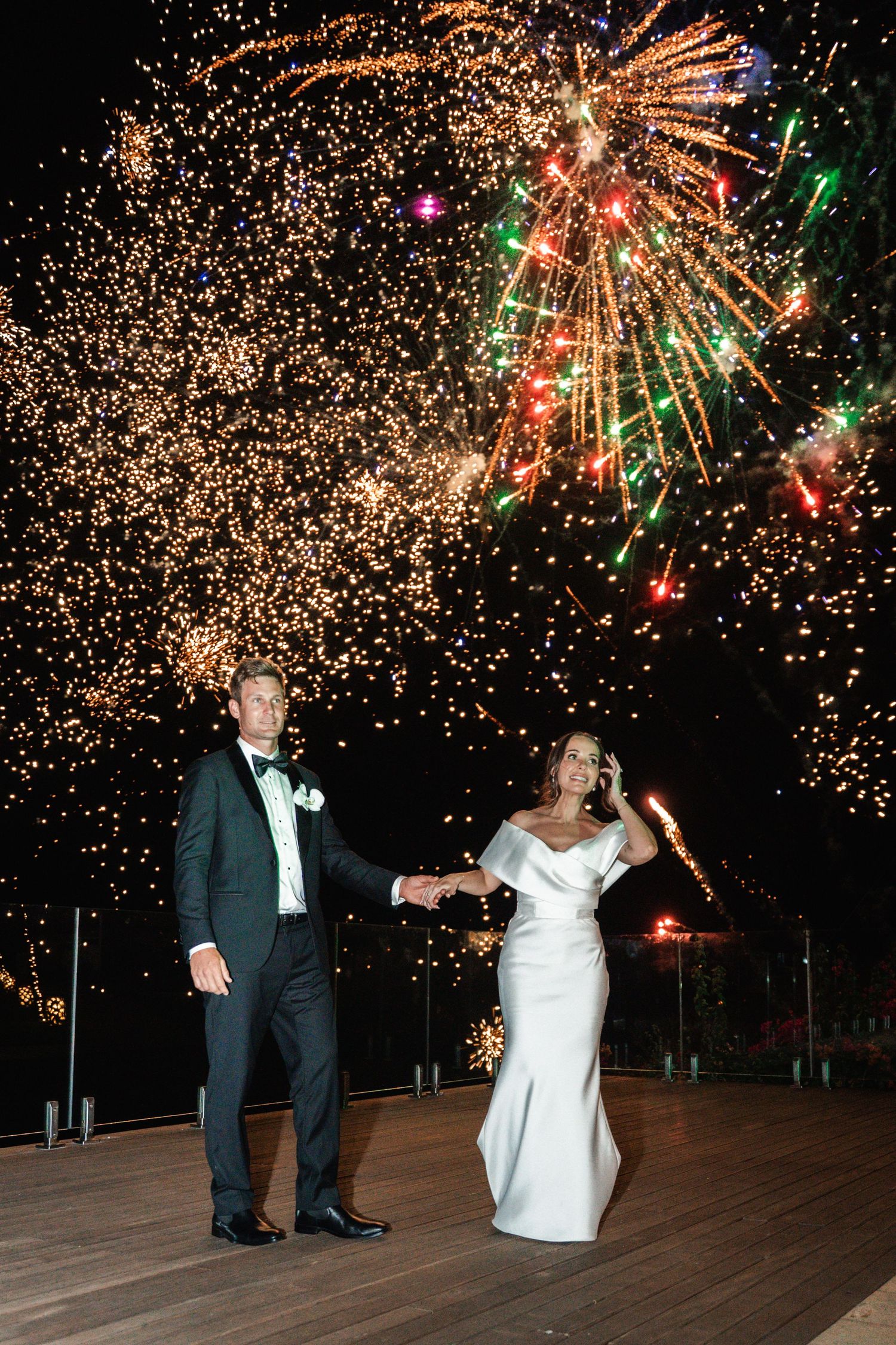 A couple celebrates their wedding reception on an outdoor dance floor while colorful fireworks explode in the night sky above.