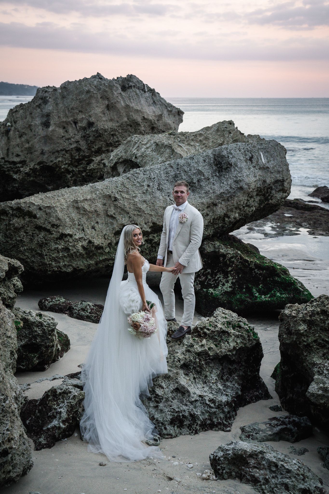 Wedding couple posing on rocky beach cliffs during sunset.