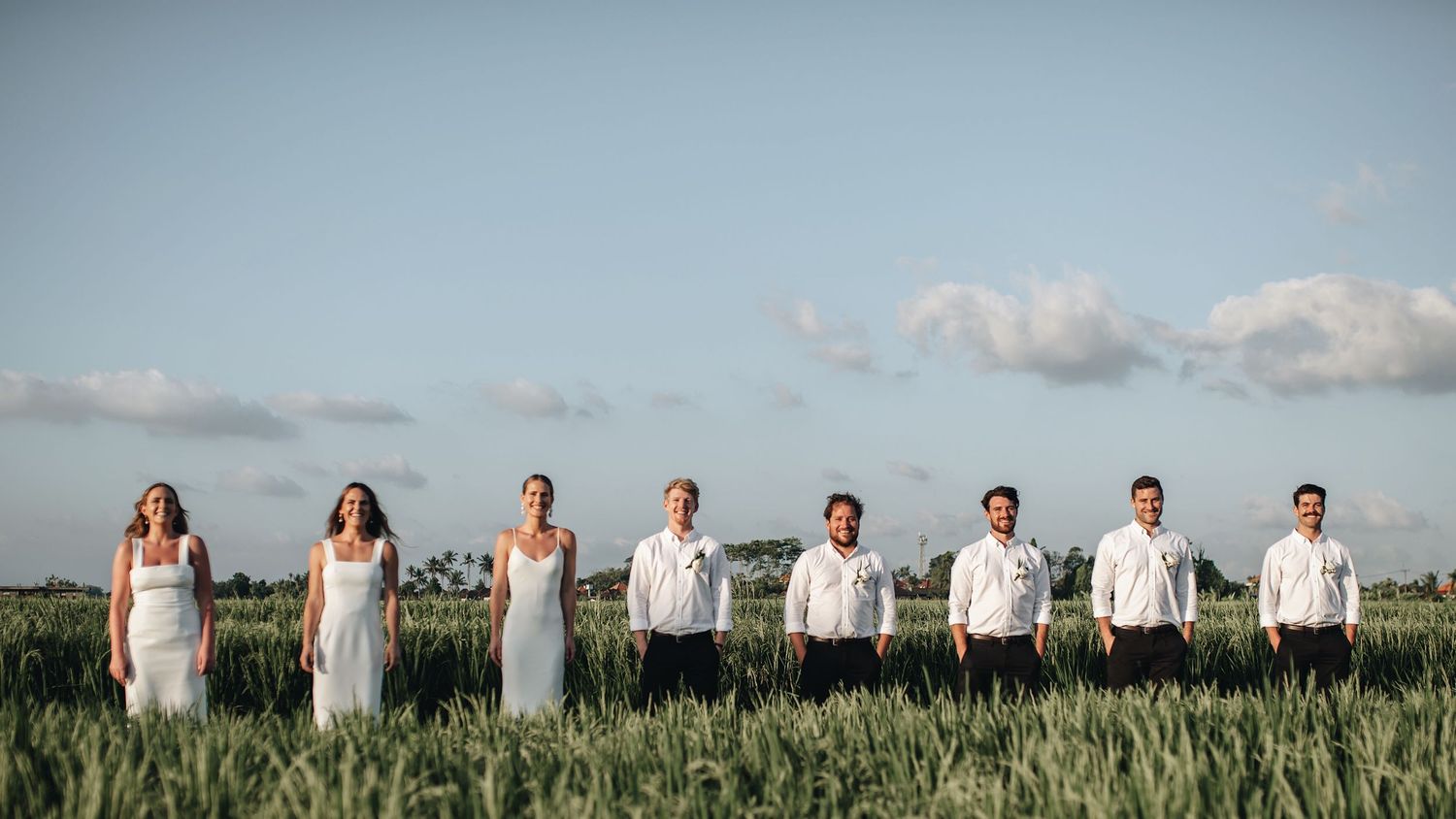 Wedding party stands in line against blue sky in grassy field.