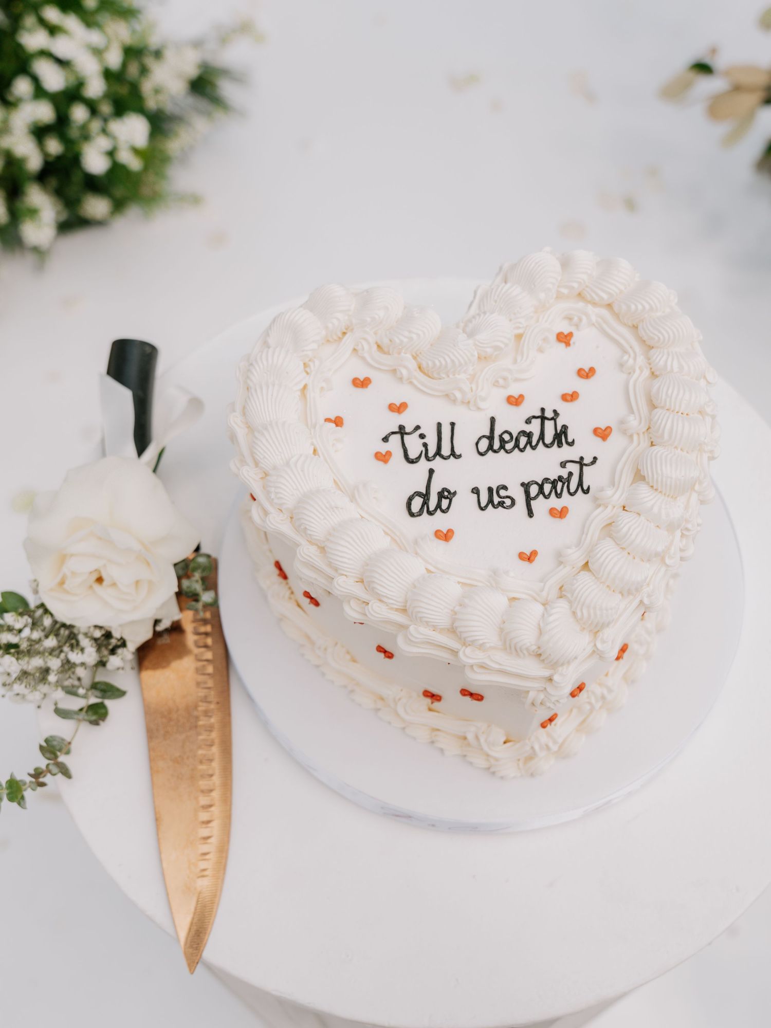 Heart-shaped white wedding cake with 'till death do us part' inscription, small hearts, and gold knife beside white roses.