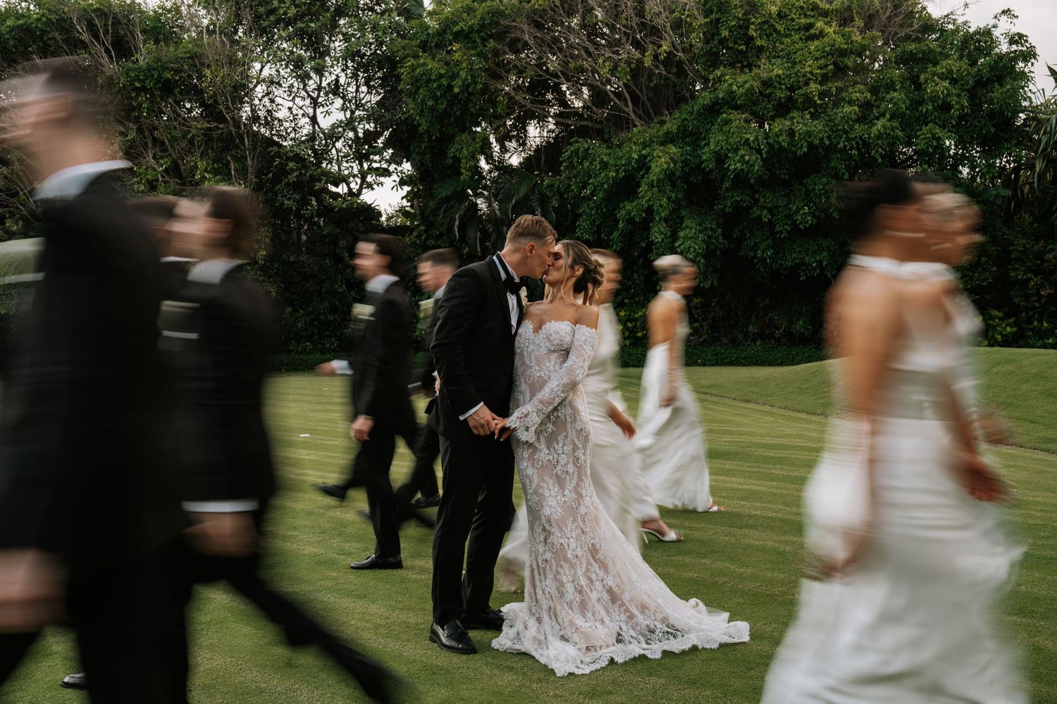 A wedding party poses together outdoors in a garden setting with a white dress flowing in the breeze.