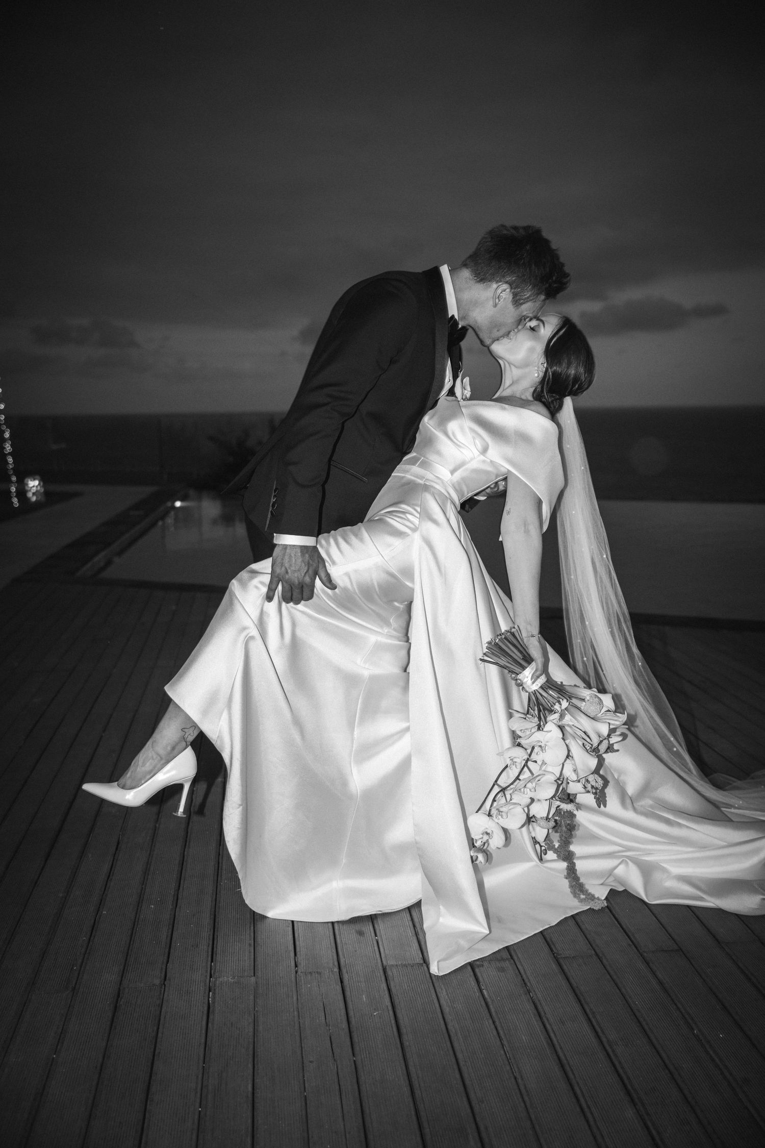 A romantic black and white sequence of a couple in wedding attire sharing an intimate moment on a wooden deck at sunset.