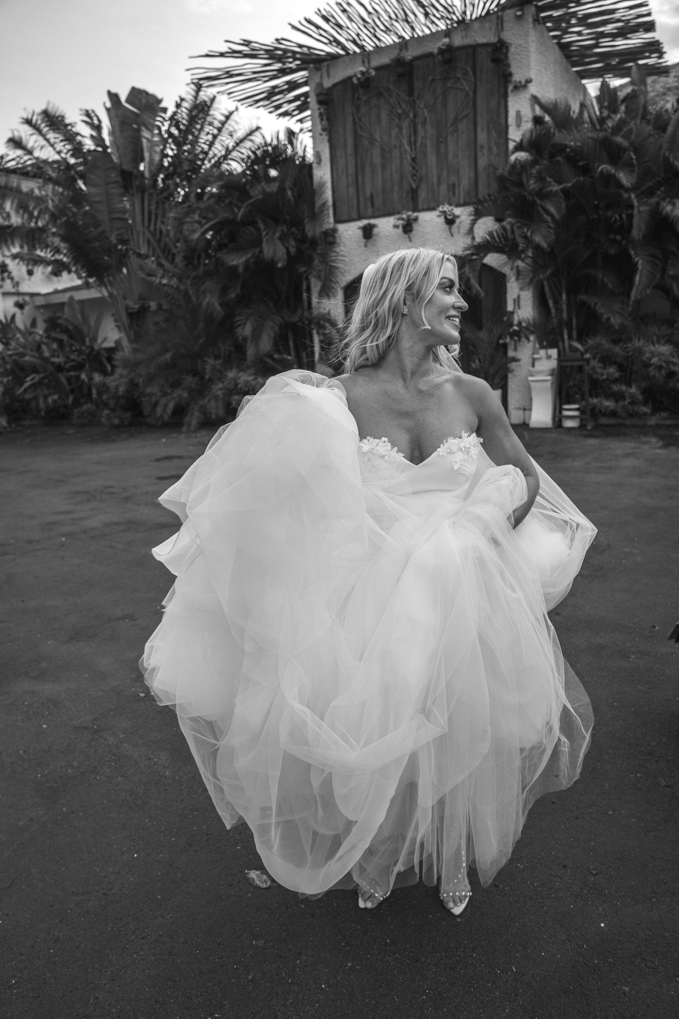 A joyful moment captured in black and white as someone in a flowing white wedding dress twirls outdoors among palm trees.