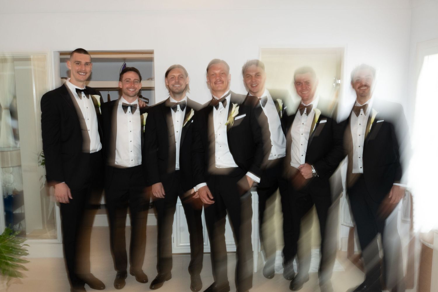A row of people in formal black tuxedos and bow ties stand together in a line against a white wall.