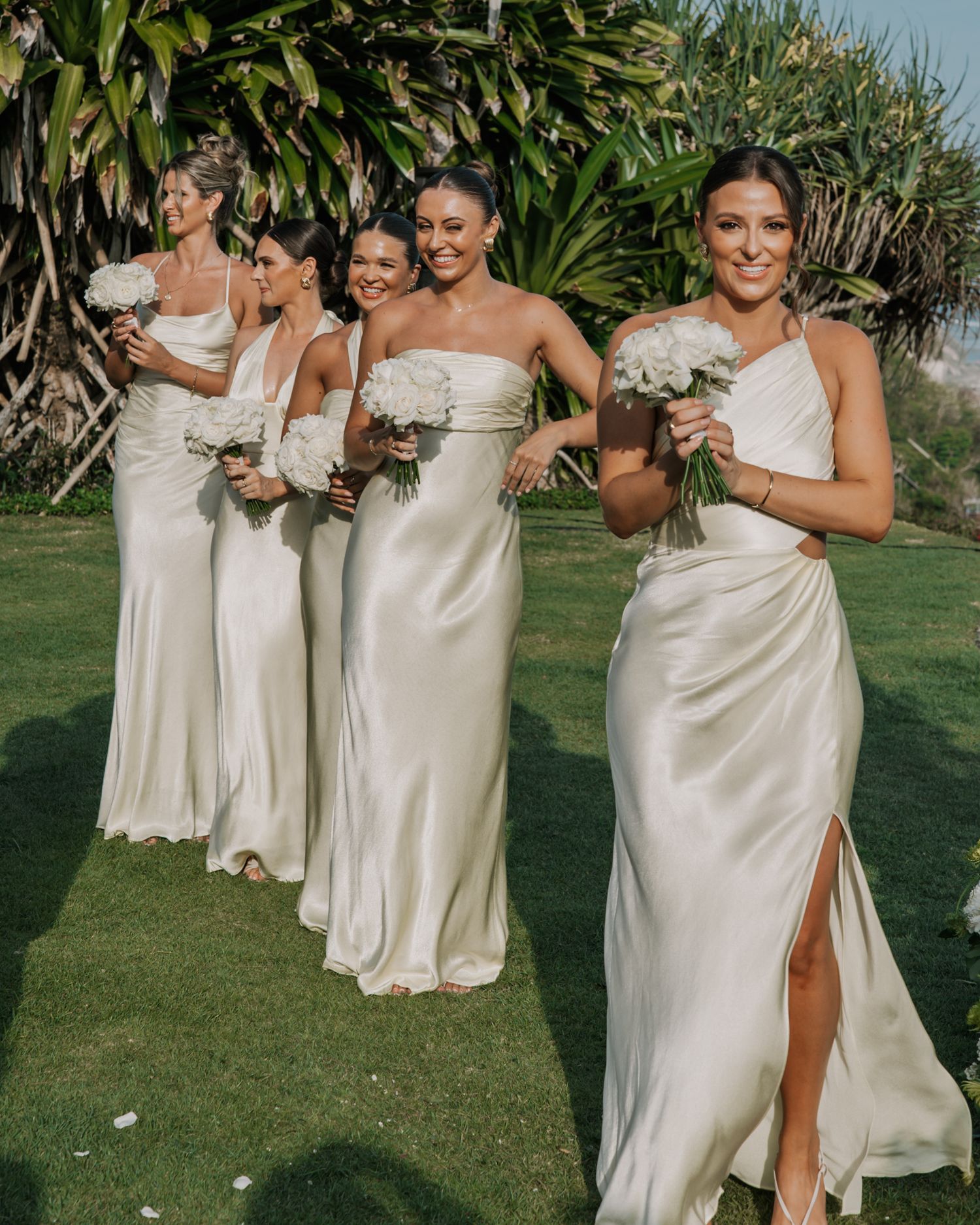 Bridesmaids in champagne silk dresses pose playfully on a sunny lawn with tropical foliage in the background.