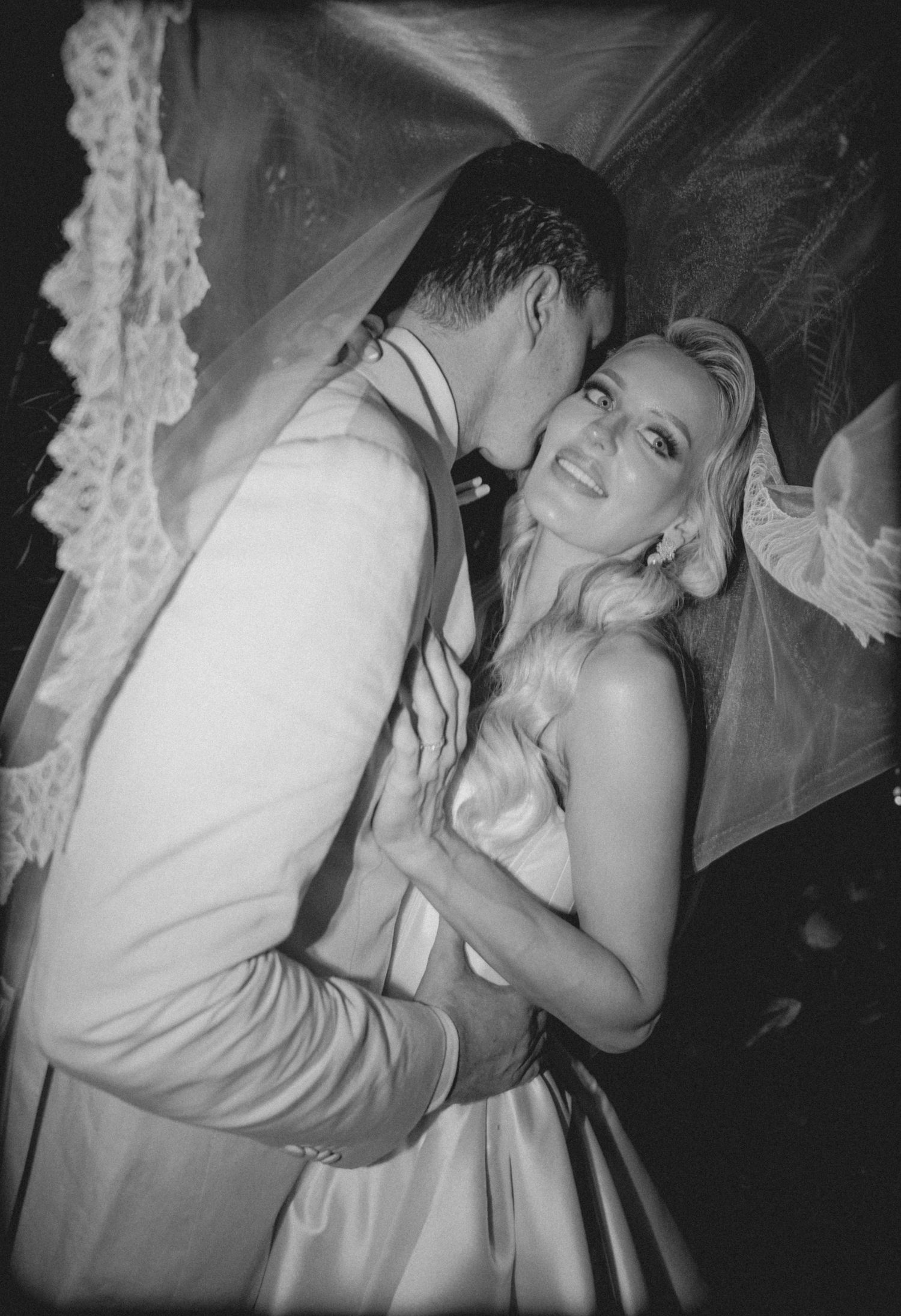 Black and white wedding photo of a groom kissing bride on cheek while she smiles, under a lace veil.